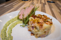 Sliced Herb-crusted Pork Tenderloin with pink center and herb crust, beside a bacon-topped potato cake.