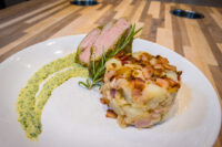 Sliced Herb-crusted Pork Tenderloin with herb crust edge, beside a potato-bacon cake and herb sauce swirl