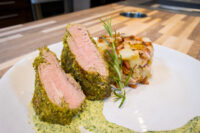 Sliced Herb-crusted Pork Tenderloin with pink centre in green herb sauce, beside a potato bake