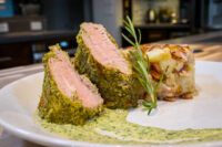 Sliced Herb-crusted Pork Tenderloin with pink centres, plated beside a potato bake and green herb sauce