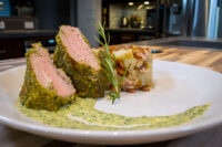 Sliced Herb-crusted Pork Tenderloin with pink center, beside a golden potato bake and green herb sauce.