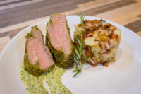 Sliced Herb-crusted Pork Tenderloin with pink centre, green herb sauce, potatoes and bacon, rosemary sprig