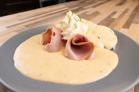 Pale Horseradish Sauce Křenová Omáčka pooled on a gray plate under rolled ham slices and whipped cream.