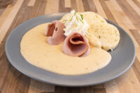 Pale creamy Horseradish Sauce pooled on a plate with rolled ham and a dumpling slice topped with whipped cream.
