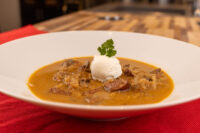 Bowl of Kapustnica Soup with orange broth, meat chunks, a cream dollop, and parsley garnish on red cloth.