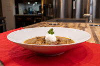 White bowl of Kapustnica Soup with a cream dollop and parsley sprig on a red tablecloth.