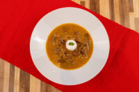 Overhead shot of Kapustnica Soup in a white bowl, topped with cream and a sprig of herb, on a red cloth.
