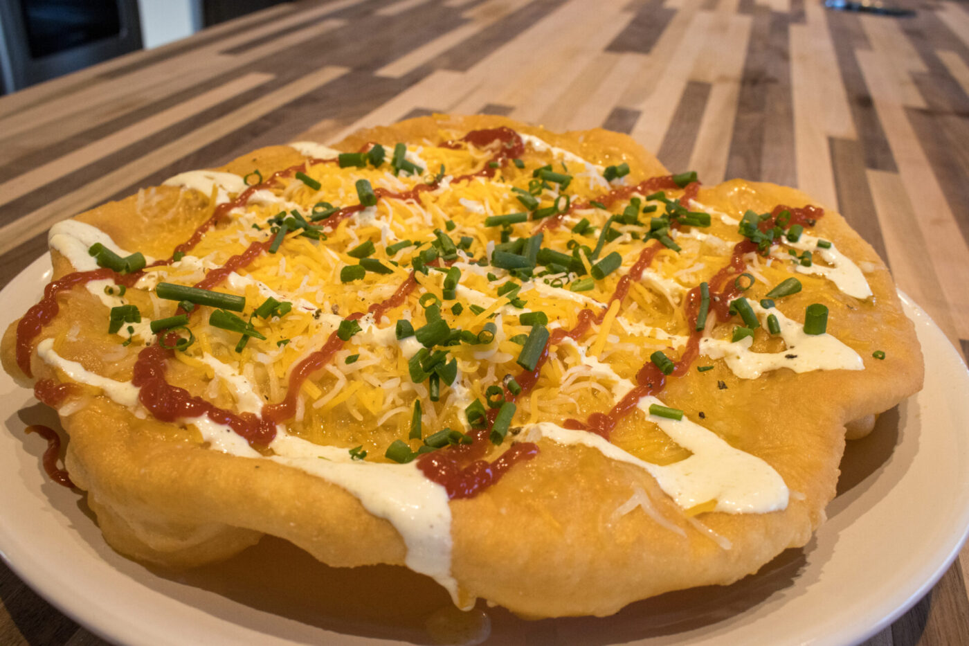 Langos flatbread on a white plate topped with sour cream, red sauce, shredded cheese, and chives.