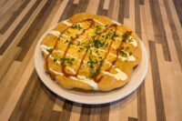 Round fried Langos flatbread topped with cheese, white and red drizzle, and chives on a white plate