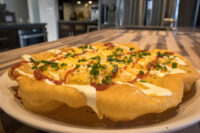 Langos: puffed fried dough topped with melted cheese, red sauce, white sauce drizzle, and chopped chives on a plate.