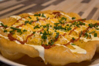Fried Langos flatbread topped with sour cream, red sauce, shredded cheese, and chives on a white plate.