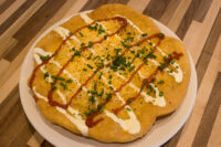 Golden fried Langos flatbread topped with cheese, chives, and red and white sauce drizzles on a plate.