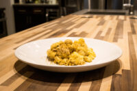 White plate of Mac & Cheese with creamy sauce and golden herb crust on a wooden counter.