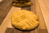 A Peanut Butter Cookie with crosshatch fork marks on a wooden board, more cookies stacked behind it.