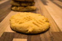 A golden Peanut Butter Cookie with crosshatch fork marks, more cookies stacked behind it on a wooden table.