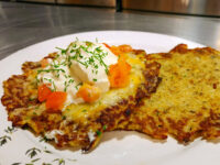 Two crisp Potato Pancakes Bramboraky topped with sour cream, diced tomato, melted cheese, and chives on a white plate.