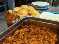 Pan of shredded pulled pork with a jar of BBQ sauce and biscuits on a cooling rack nearby.