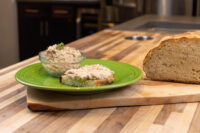Bowl of Sardines Dip or Spread beside a slice of bread topped with the spread, on a green plate with a loaf nearby.