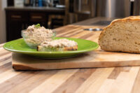 Glass bowl of Sardines Dip or Spread beside a slice topped with it, with a crusty loaf on a wood board.