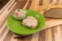 Glass bowl of Sardines Dip or Spread beside a slice of bread topped with it, with a crusty loaf on a wooden board