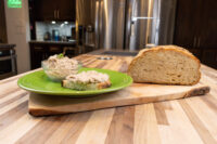 Green plate with bowl of Sardines Dip or Spread and a topped bread slice, beside a whole loaf of bread on a wooden board
