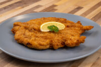 Chicken Schnitzel Golden fried Chicken Schnitzel on a grey plate, topped with a lemon wedge and parsley sprig.