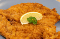 Chicken Schnitzel Golden breaded Chicken Schnitzel on a gray plate with a lemon wedge and parsley garnish.