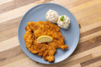 Chicken Schnitzel Golden breaded Chicken Schnitzel with a lemon wedge, served alongside creamy coleslaw on a gray plate.