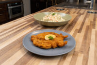 Chicken Schnitzel Golden fried chicken schnitzel with lemon slice on a grey plate, next to a bowl of creamy salad.