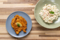 Chicken Schnitzel Golden Chicken Schnitzel with a lemon wedge on a grey plate beside a bowl of potato salad.