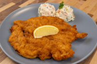 Chicken Schnitzel Golden fried Chicken Schnitzel topped with a lemon wedge, served on a gray plate with potato salad.