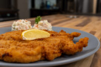 Chicken Schnitzel Golden fried Chicken Schnitzel on a grey plate with a lemon slice and creamy salad garnish.