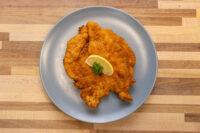 Chicken Schnitzel Golden breaded Chicken Schnitzel on a grey plate, garnished with a lemon slice and fresh herb sprig.