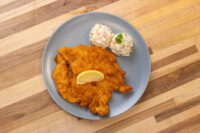Chicken Schnitzel Golden breaded Chicken Schnitzel on a grey plate with a lemon wedge and creamy potato salad.