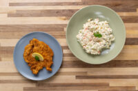 Chicken Schnitzel Golden Chicken Schnitzel with lemon wedge on a grey plate beside creamy potato salad on wood table.