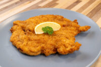 Chicken Schnitzel Golden fried Chicken Schnitzel on a grey plate, topped with a lemon wedge and parsley garnish.