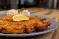 Chicken Schnitzel Golden breaded Chicken Schnitzel on a grey plate with a lemon wedge and creamy side salad.