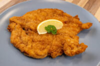 Chicken Schnitzel Golden fried Chicken Schnitzel on a grey plate, garnished with a lemon slice and parsley.