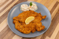 Chicken Schnitzel Golden breaded Chicken Schnitzel on a grey plate with a lemon wedge and creamy potato salad.