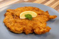 Chicken Schnitzel Golden-fried Chicken Schnitzel on a grey plate, garnished with a lemon wedge and parsley sprig.