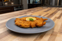 Chicken Schnitzel Golden fried Chicken Schnitzel on a gray plate with a lemon slice and herb garnish on a wooden counter.