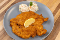Chicken Schnitzel Golden breaded Chicken Schnitzel with a lemon wedge, served beside creamy potato salad on a gray plate.