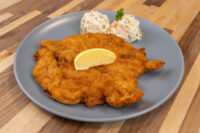 Chicken Schnitzel Golden fried Chicken Schnitzel on a grey plate with a lemon wedge and two scoops of creamy salad.