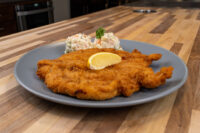 Chicken Schnitzel Golden breaded Chicken Schnitzel on a grey plate with a lemon wedge and potato salad garnished with parsley.