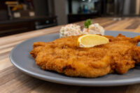 Chicken Schnitzel Golden fried Chicken Schnitzel topped with a lemon wedge, served with a creamy salad on a grey plate.