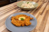 Chicken Schnitzel Golden breaded Chicken Schnitzel on a grey plate with lemon wedge and herb, beside a bowl of creamy salad.