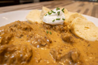 Bowl of Szegedin Goulash with creamy paprika sauce, sour cream, chives, and bread slices on a white plate.