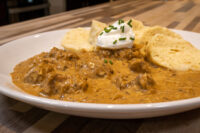 Bowl of Szegedin Goulash with creamy paprika sauce, sour cream, chives, and bread slices on a white plate.