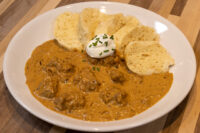 Bowl of Szegedin Goulash with meat in creamy paprika sauce, sour cream, chives, and bread rolls.