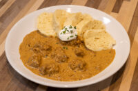 Bowl of Szegedin Goulash with sour cream and chives, served with sliced bread on a wooden table.